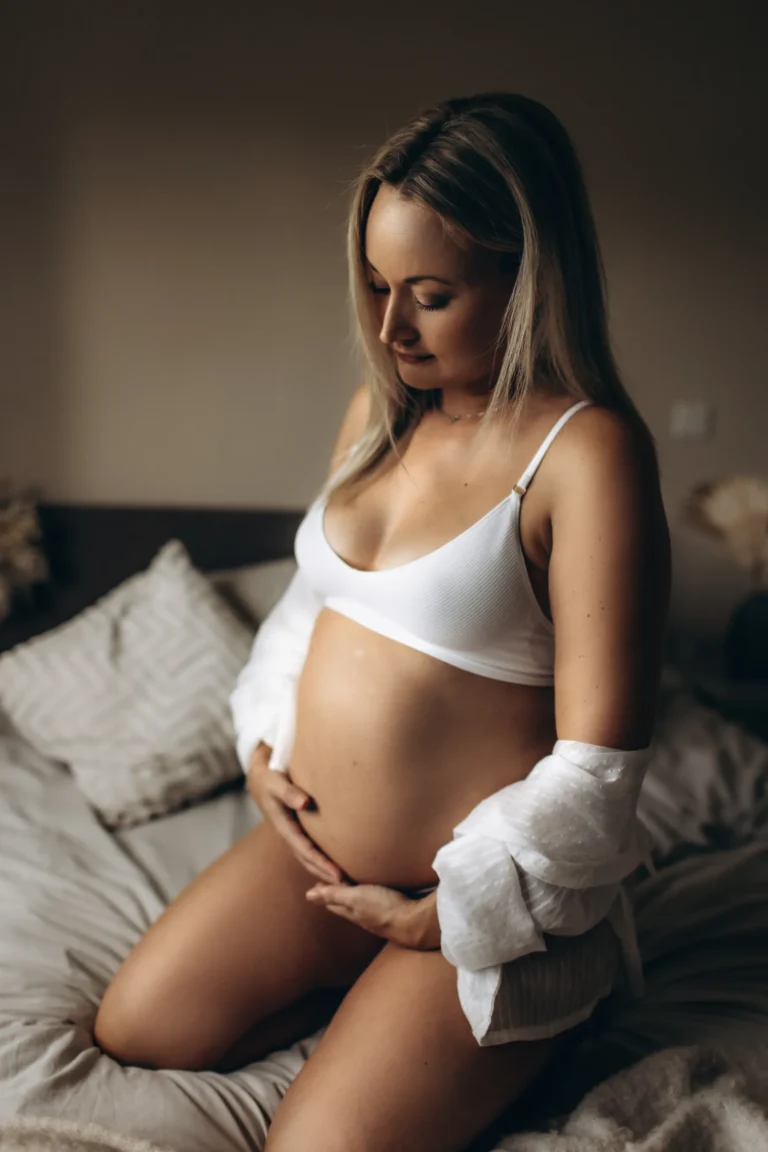 A pregnant woman with long blonde hair, wearing a white bra and a partially open white shirt, sits on a bed with neutral-toned bedding during her London in-home maternity photo session. She gently cradles her belly, looking down with a peaceful expression.