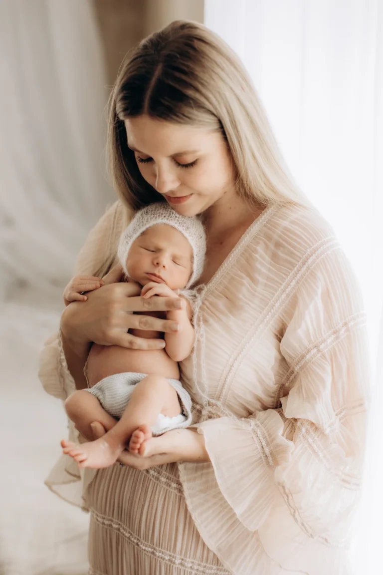 A woman wearing a sheer, light-colored dress cradles a sleeping baby wrapped in a white knit hat and diaper. She gazes lovingly at the infant, who is held close to her chest. The background is softly lit, creating a serene and intimate atmosphere.