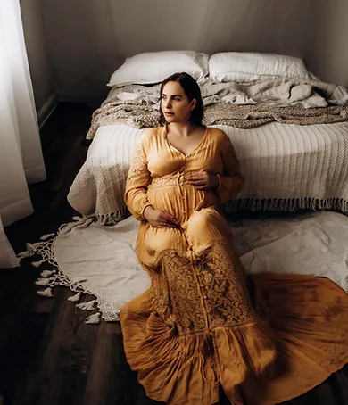 A pregnant woman in a long, flowing yellow dress sits on a white rug in front of a bed. She is holding her belly and looking off to the side, clearly captured by a talented pregnancy photographer. The room has a cozy, calm ambiance with soft lighting, a white bedspread, and pillows neatly arranged.