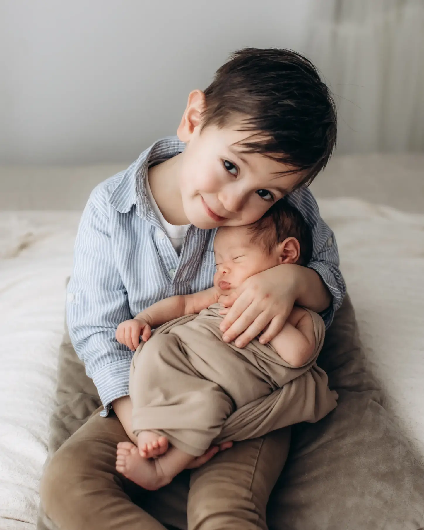 A young boy with dark hair, wearing a blue striped shirt and brown pants, sits on a bed and gently holds a sleeping newborn baby wrapped in a beige blanket. Both children appear peaceful and content in a cozy setting captured beautifully by a Surrey family photographer.