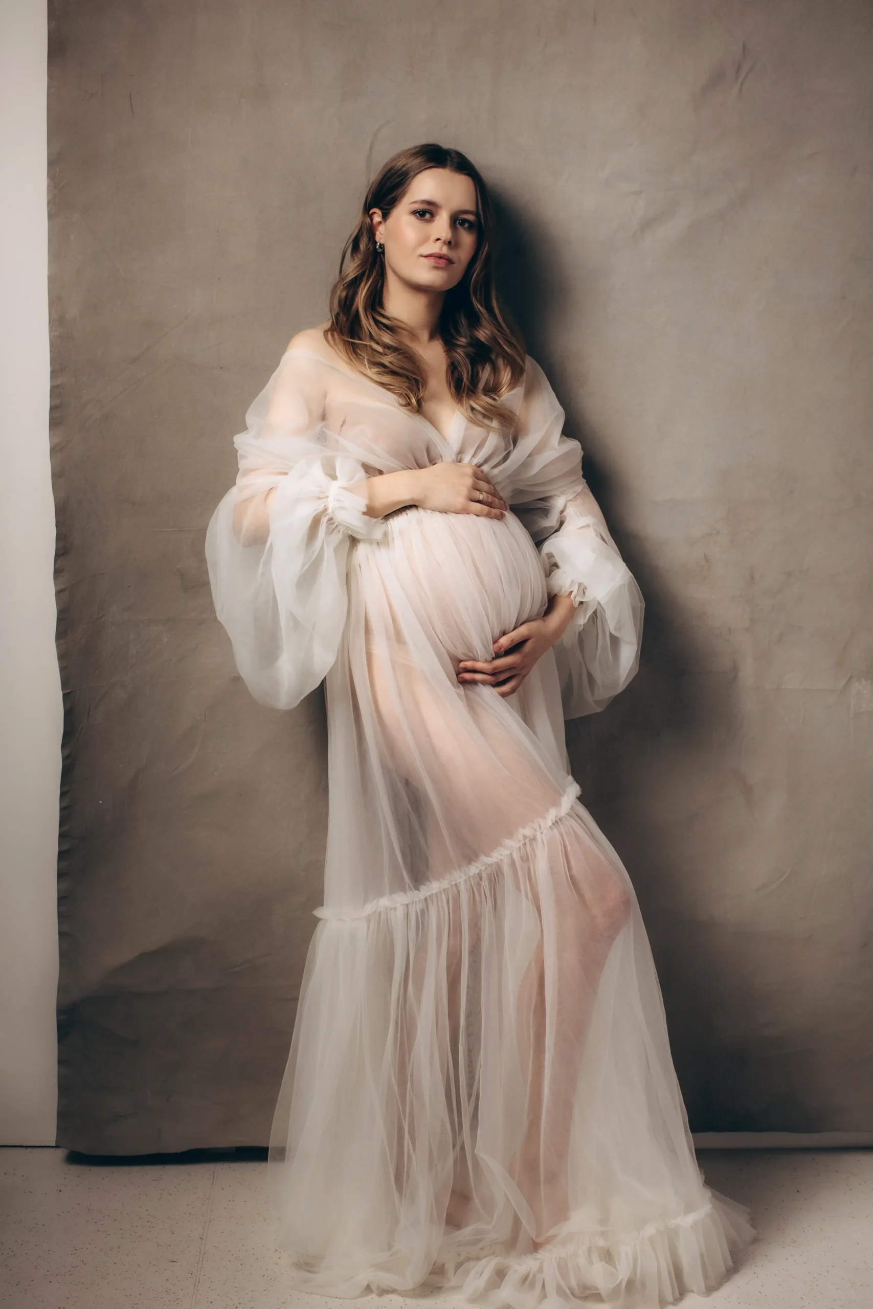 A pregnancy photographer captures a pregnant person standing against a gray backdrop, gently holding their belly with both hands. They wear a sheer, flowing white dress with long, puffed sleeves and have long, wavy hair cascading over their shoulders. The atmosphere is serene and elegant.