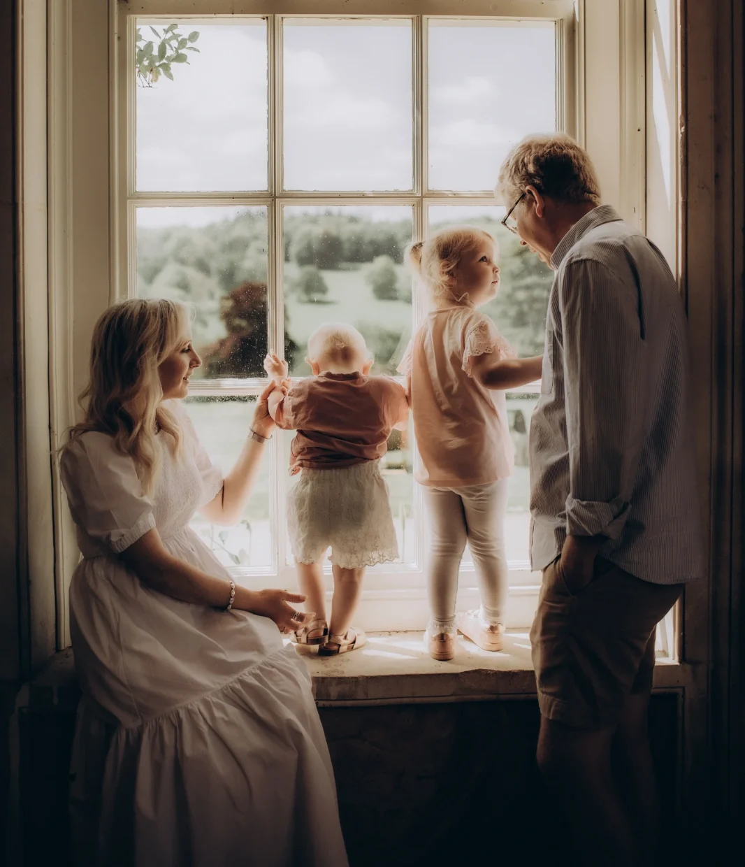 A family of four is gathered by a large window. A woman in a white dress sits on the left, while a man in casual clothes stands on the right. Two young children, one standing and one being held, look out the window at the scenic landscape outside, capturing a perfect moment of Surrey Newborn Photography.