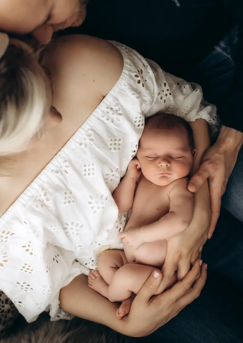 A newborn baby sleeps peacefully in the arms of a person wearing a white, off-shoulder blouse. The baby is swaddled in light clothing, and another person gently cradles them from above, creating a warm, protective embrace — beautifully captured by Surrey Newborn Photography.