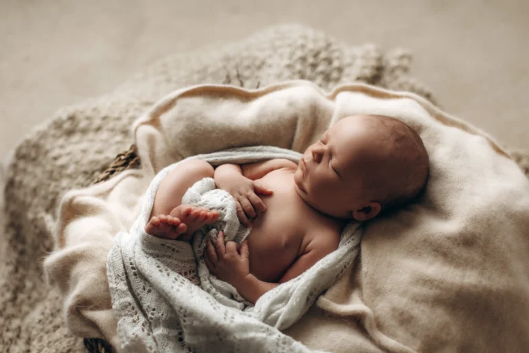 A newborn baby sleeps peacefully on its back, nestled in soft cream-coloured blankets. The baby is swathed in a light lace cloth, with one hand resting on the chest and small feet peeking out. The background is out of focus but appears cozy and warm. Lifestyle newborn photography session in Surrey