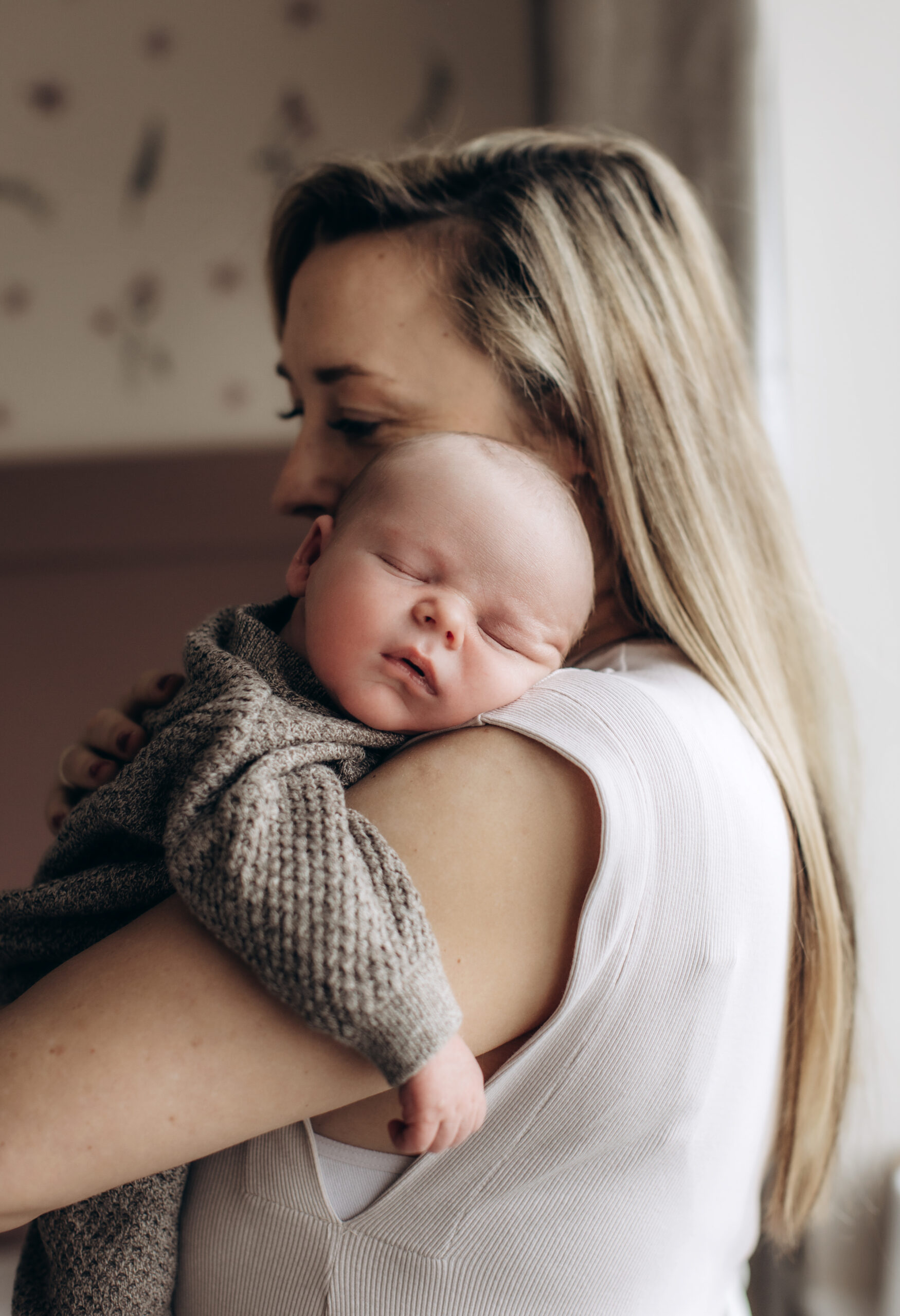 A woman with long blonde hair is holding a sleeping baby on her shoulder. The baby is dressed in a knitted outfit and appears peaceful. The background is softly blurred, featuring warm tones and subtle patterns. The scene evokes a feeling of warmth and tenderness.