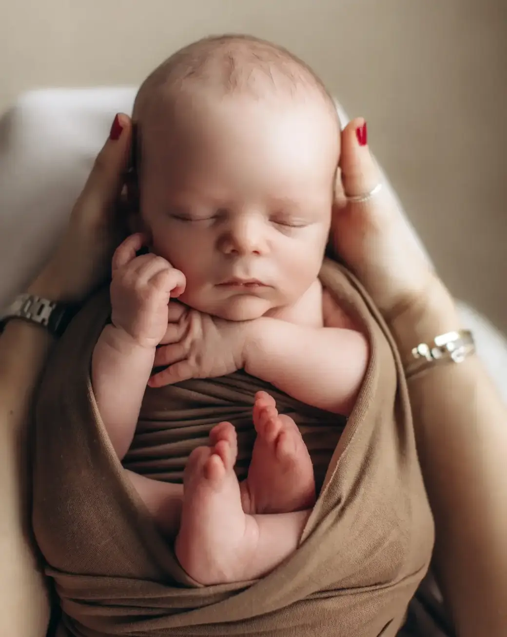 A peaceful baby wrapped in a brown blanket is being cradled by adult hands. The baby is asleep with hands near the face and small feet peeking out. The adult's hands, with red-painted nails, gently hold the baby, creating a sense of warmth and protecti...