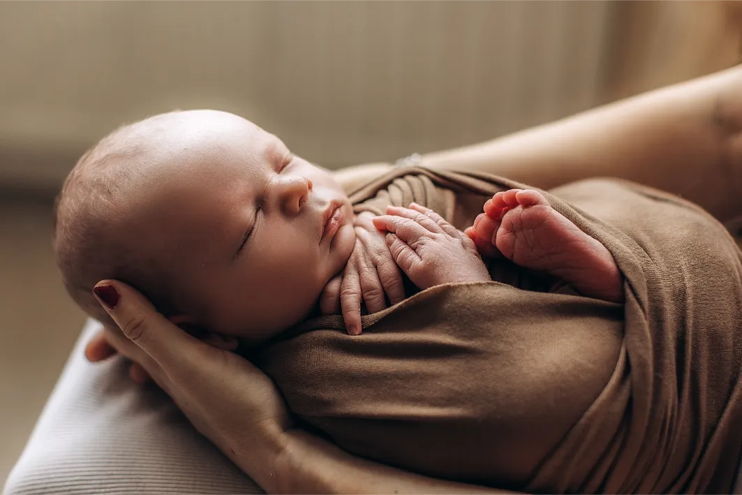 Close-up of a peacefully sleeping newborn baby wrapped in a brown blanket, being gently cradled in an adult's arms. The baby's hands are tucked under their chin, and their tiny feet are visible. The scene exudes warmth and tranquility, capturing the essence of lifestyle newborn photography in Surrey.