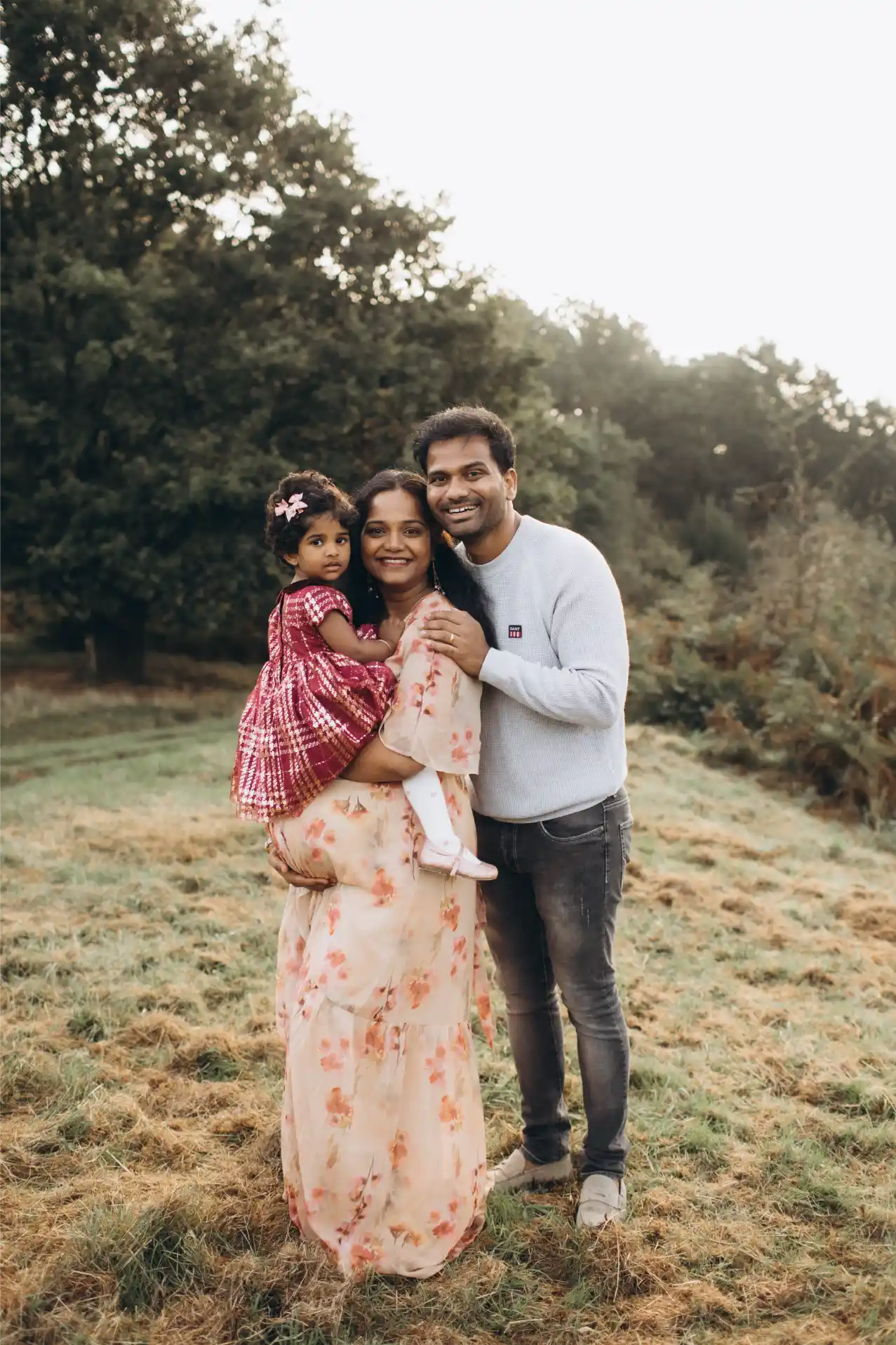 A family of three poses outdoors in a grassy area with trees in the background, captured beautifully by a family photographer in Surrey. The woman holds a young child dressed in a red checkered outfit, wearing a floral dress herself. The man stands beside them, smiling in his light gray sweater and jeans.