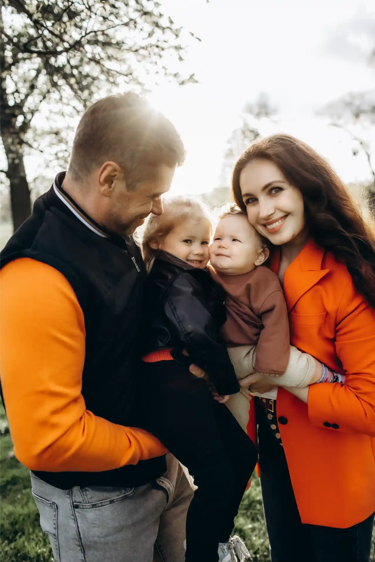 A cheerful family of four stands outdoors. The father, dressed in an orange and black jacket, holds a small girl in a black jacket, while the mother, in an orange coat, cradles a baby. They all smile warmly, with sunlight filtering through the trees in the background. Captured perfectly by their family photographer in Surrey.