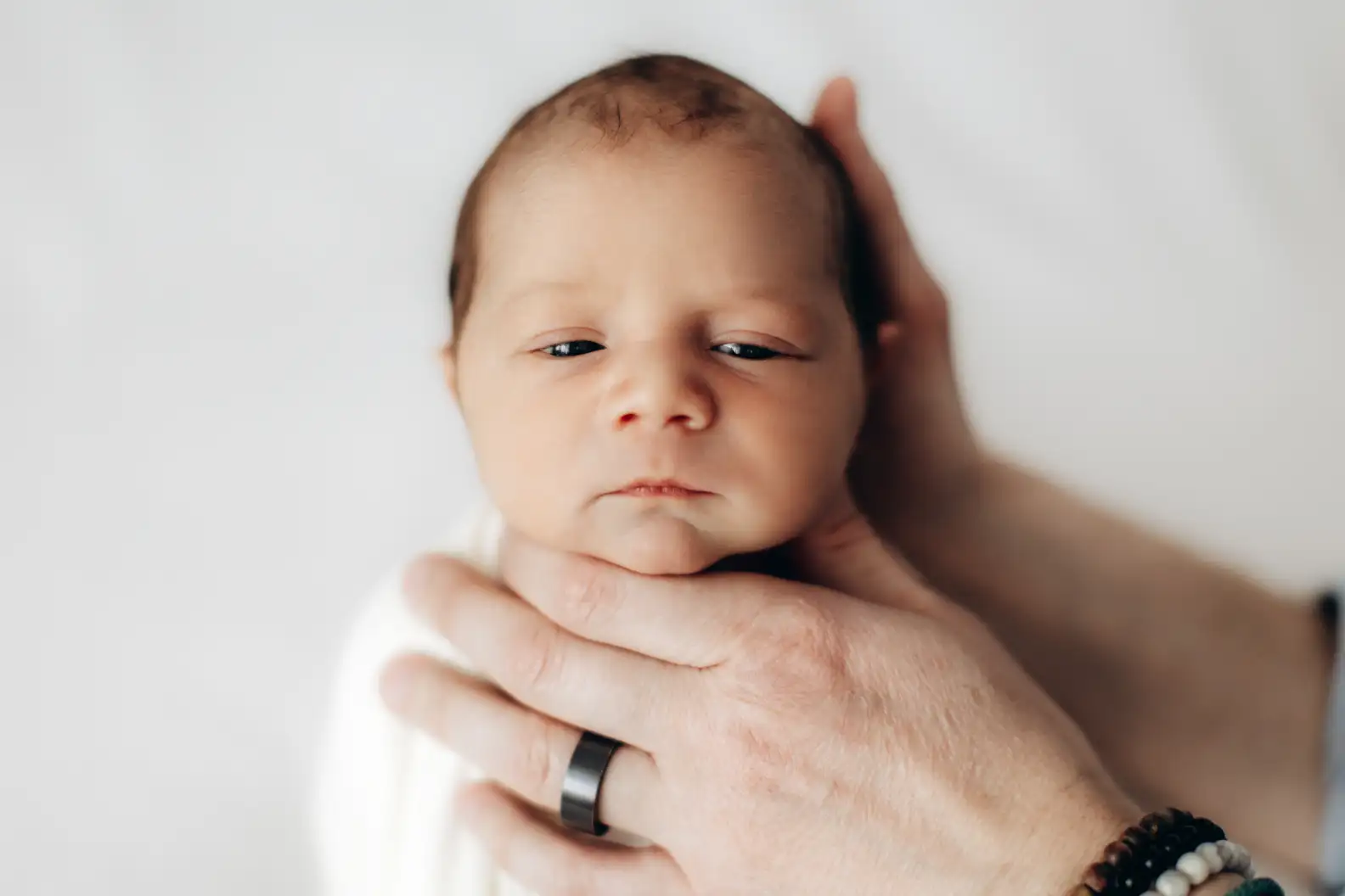 A newborn baby, photographed by a top newborn photographer, is swaddled in a white blanket and gently cradled by adult hands. The baby has closed eyes and a calm expression. The adult wears a dark ring on one finger and a bracelet made of beads. The background is soft and white.