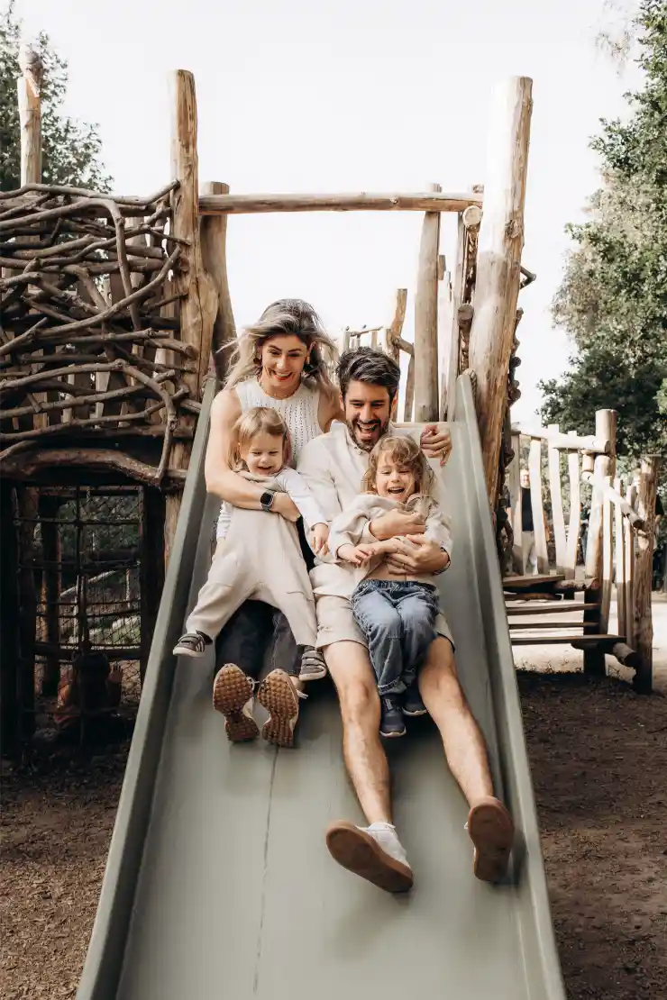 A happy family of four goes down a playground slide together. The parents, a father and mother, sit behind their two young children, all smiling and enjoying the ride. Captured by a Surrey family photographer, the wooden playground structure and trees are visible in the background.