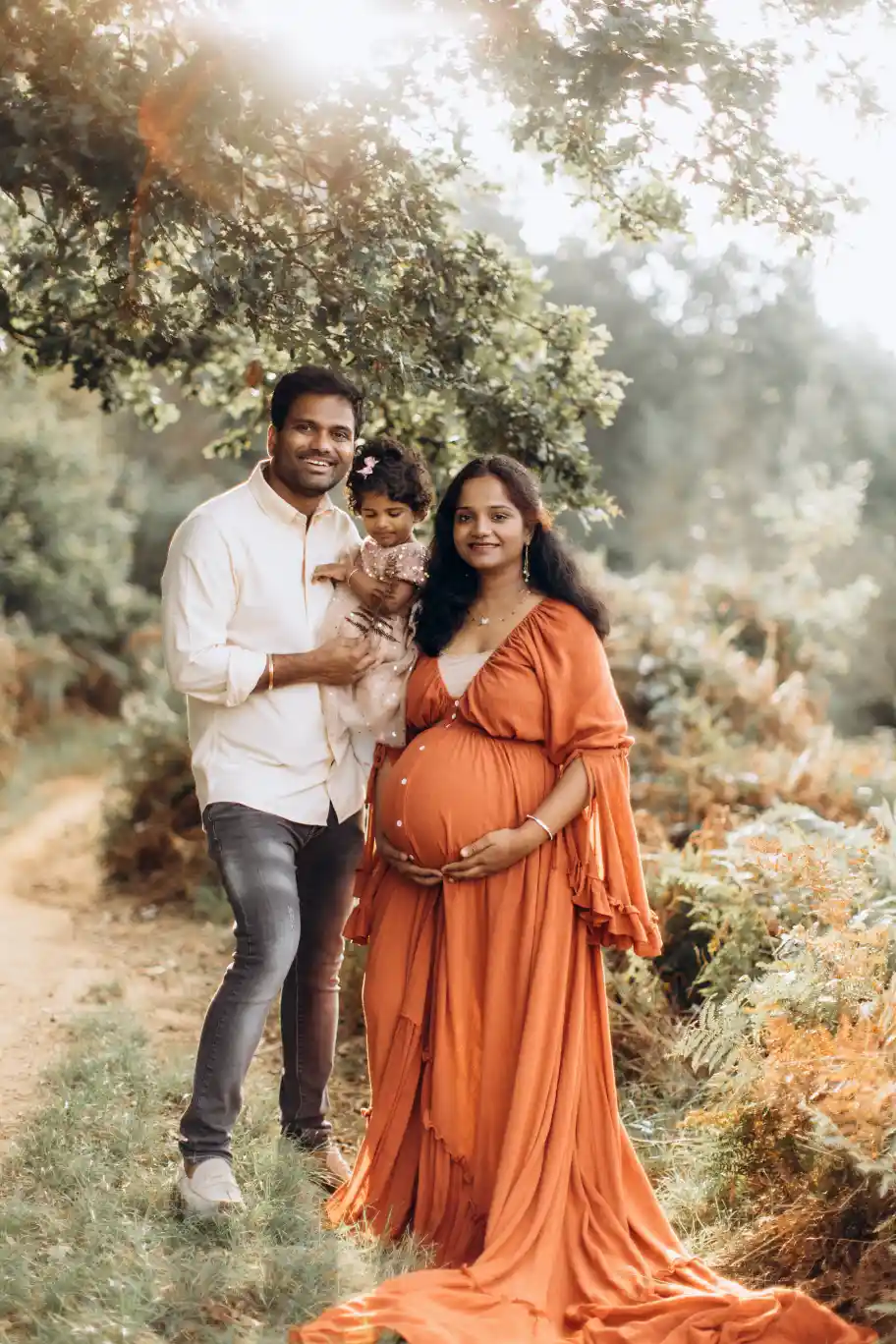 A smiling family stands outdoors on a wooded path. The father, in a white shirt, holds a small child wearing a beige outfit. The pregnant mother, in a flowing orange dress, rests one hand on her baby bump. Captured by Surrey Family Photographer, they stand beneath a tree with greenery in the background.