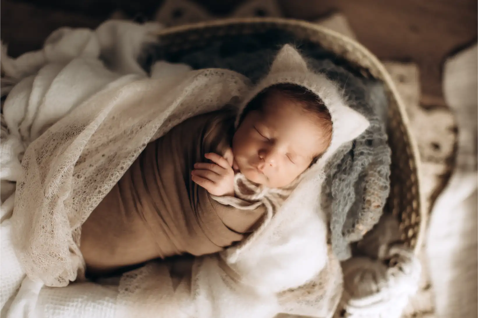 A newborn baby is sleeping peacefully, wrapped in a soft brown blanket with delicate, white lace details. The baby is wearing a cute, fuzzy hat with tiny ears, resting on a cozy bed of fabric in a neutral-toned setting—captured by the best newborn photographer.