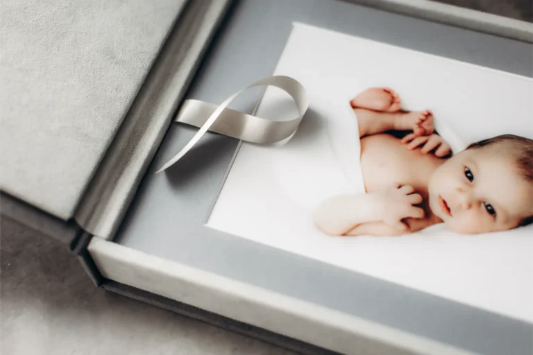 A photo album lies open, displaying a picture from a Surrey Newborn Photographer of a baby swaddled in a white blanket. The album features a grey cover with a satin ribbon bookmark resting on the page.