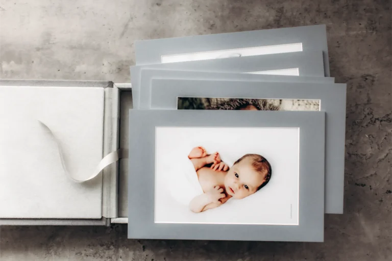 A set of photo frames with grey borders and pictures from a newborn photoshoot, partially inside an open grey box with a white ribbon, placed on a textured grey surface. The baby in the visible photo is lying on a white background, capturing precious moments from the newborn photoshoot near me.