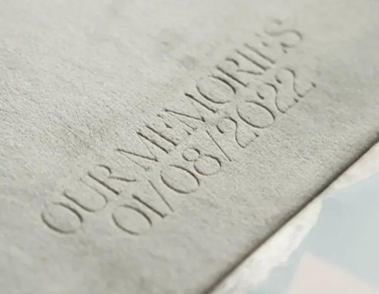 A close-up of a textured gray surface with the embossed text "OUR MEMORIES 01/08/2022." The letters and numbers appear raised and slightly faded, suggesting an etched or engraved design. A reflective surface is visible in the bottom right corner, reminiscent of a newborn photoshoot announcement.