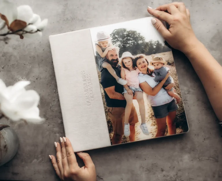 A person holding a photo album with a family picture on the cover. The beige album, embossed with "Our Memories," captures the joy of a Surrey Family Photographer's touch. The picture shows a family of five posing outdoors. A vase with white flowers graces the left side of the table.