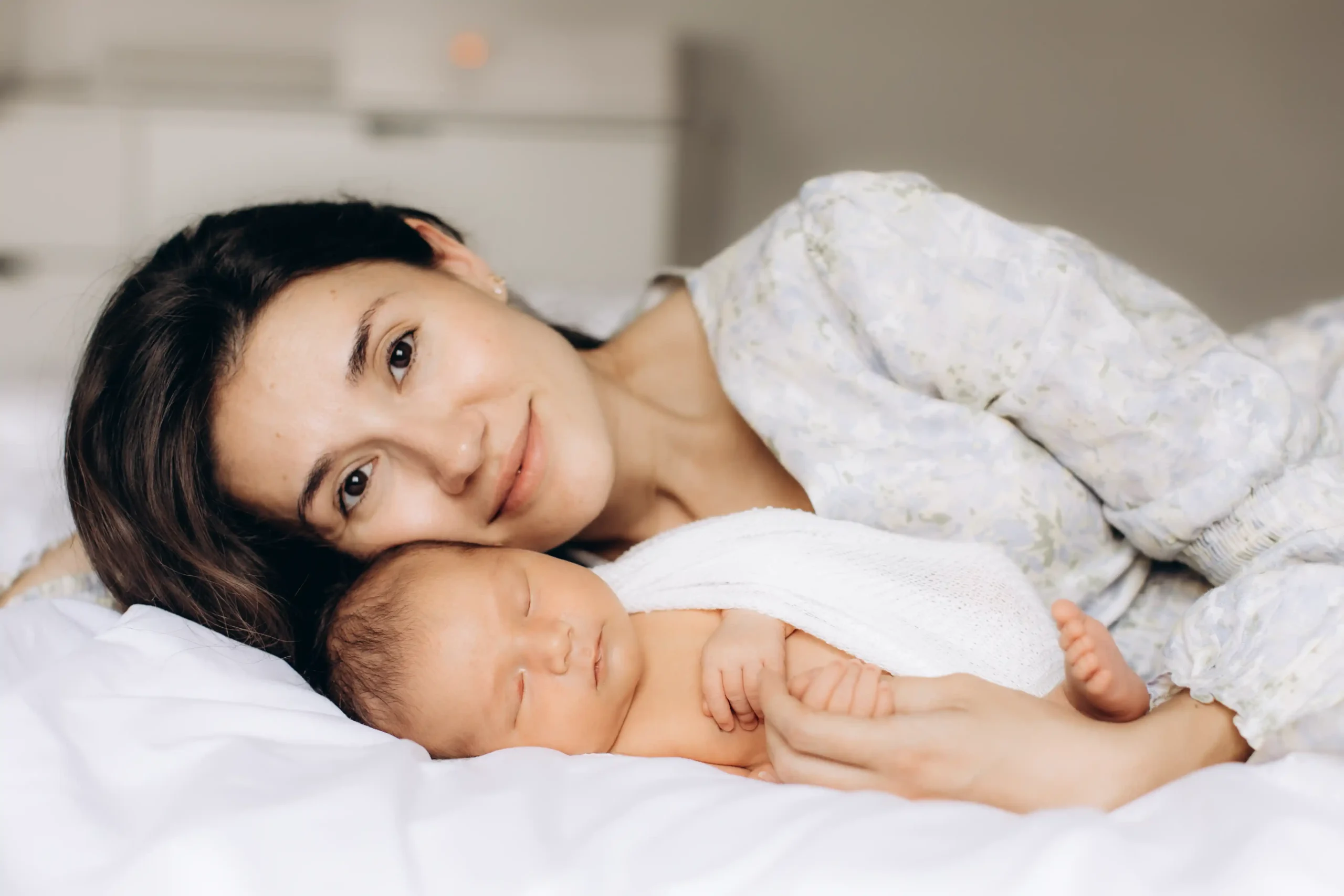 A woman lies on a white bed next to a sleeping baby swaddled in a soft, light-colored blanket. She gazes lovingly at the baby, with their faces close together. The scene is serene and tender, highlighted by soft natural light. Weybridge newborn photoshoot