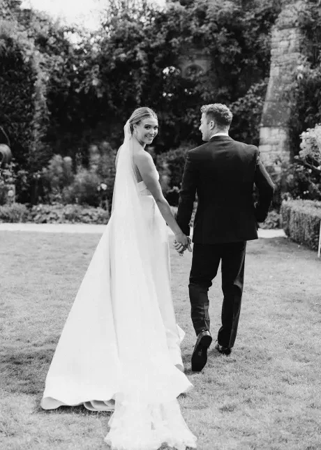 A bride and groom are walking hand in hand in a garden, captured perfectly by your Surrey wedding photographer. The bride, wearing a long white dress and veil, looks back and smiles at the camera while the groom, dressed in a dark suit, faces forward. Lush greenery and partial stone ruins adorn the background.