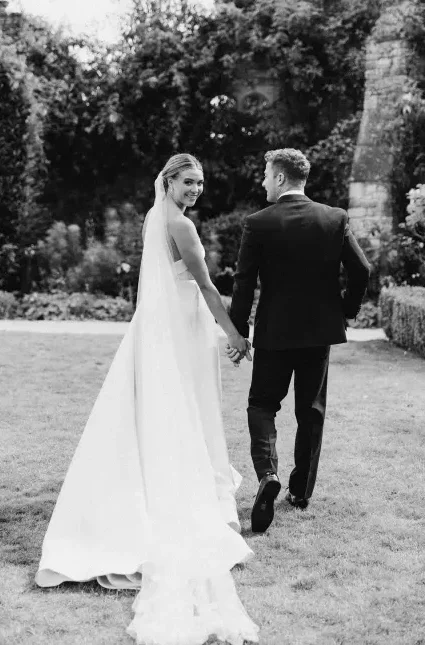 A bride and groom are walking hand in hand in a garden, captured perfectly by your Surrey wedding photographer. The bride, wearing a long white dress and veil, looks back and smiles at the camera while the groom, dressed in a dark suit, faces forward. Lush greenery and partial stone ruins adorn the background.