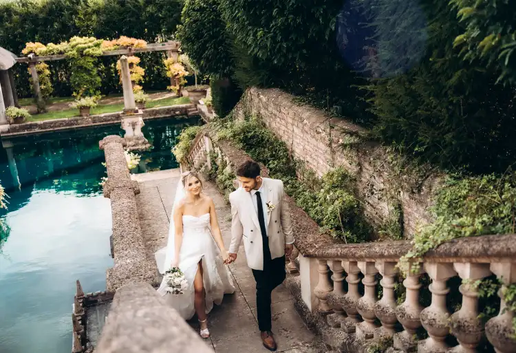 A bride and groom walk hand in hand up a stone staircase beside a serene pool surrounded by lush greenery. The bride wears a white dress and carries a bouquet, while the groom is dressed in a light-colored suit. Captured by a talented Surrey wedding photographer, the setting appears romantic and picturesque.