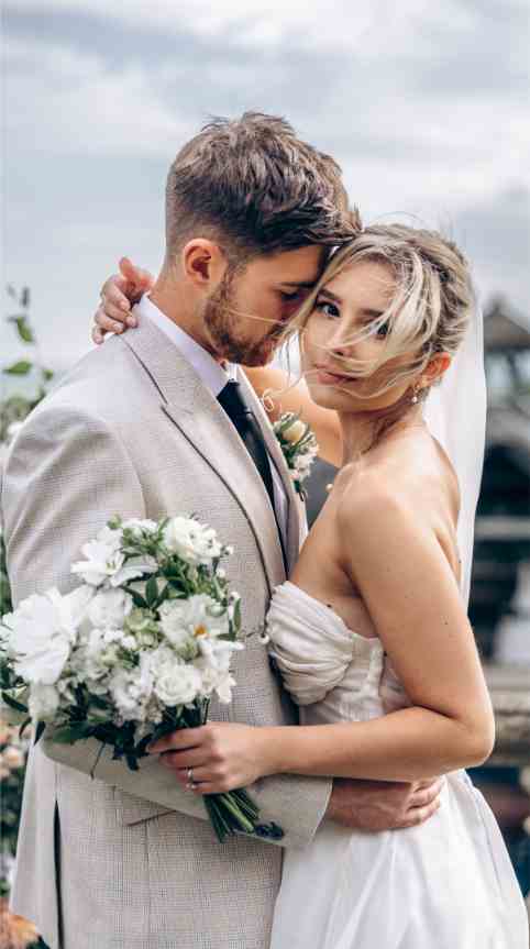 A couple in wedding attire embrace outdoors. The groom wears a light gray suit, and the bride is in a strapless white gown, holding a bouquet of white flowers. Captured by a talented Surrey family photographer, they stand close, the bride glancing at the camera with a gentle expression.