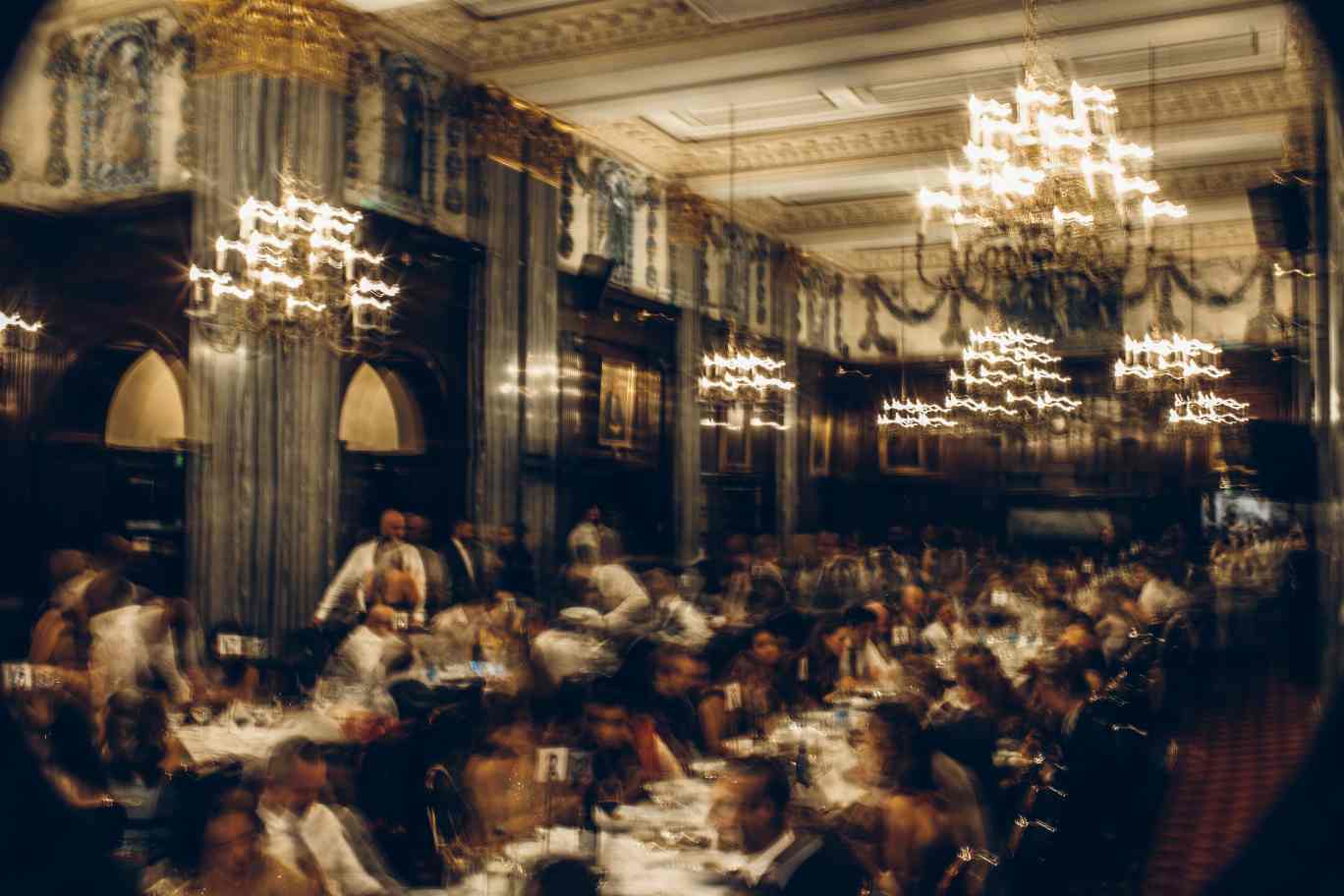 A blurred image captures the elegance of a formal banquet, possibly photographed by a Surrey wedding photographer, in an opulent room with chandeliers and ornate ceilings. Guests sit at long tables at The Law Society Hall in London with white tablecloths, engrossed in conversation. The warmly lit scene radiates liveliness and refinement.