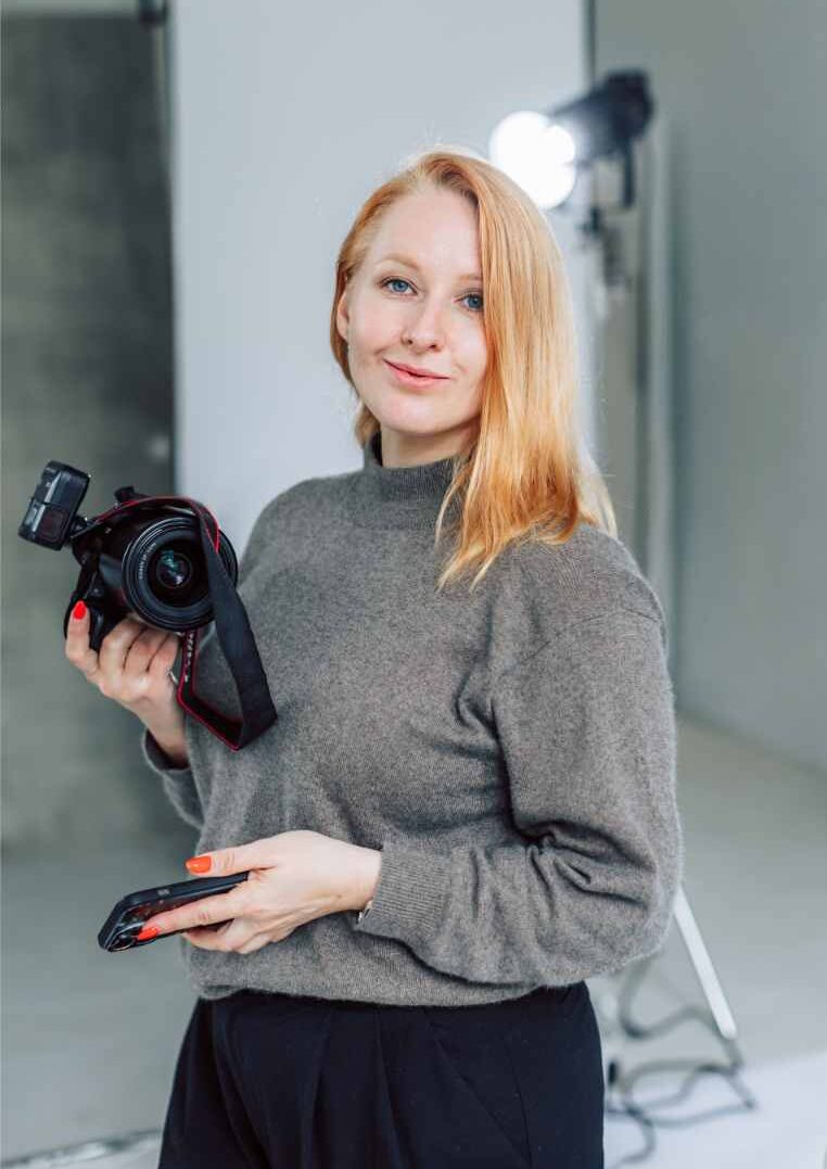 In a photography studio, a Surrey Wedding Photographer stands confidently, holding a professional camera and smartphone. Clad in a gray sweater, they are surrounded by studio lighting that enhances their creative space.