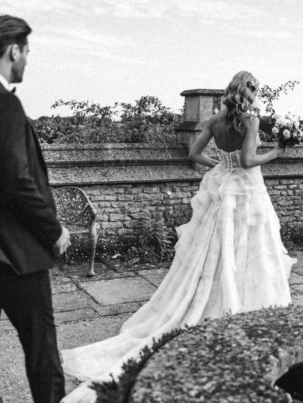 Home 2 A black and white photo captures a bride in a flowing gown holding a bouquet as she walks outdoors near a stone wall, with her groom close behind. Taken by a talented Surrey wedding photographer, the moment shines against the backdrop of a clear sky.