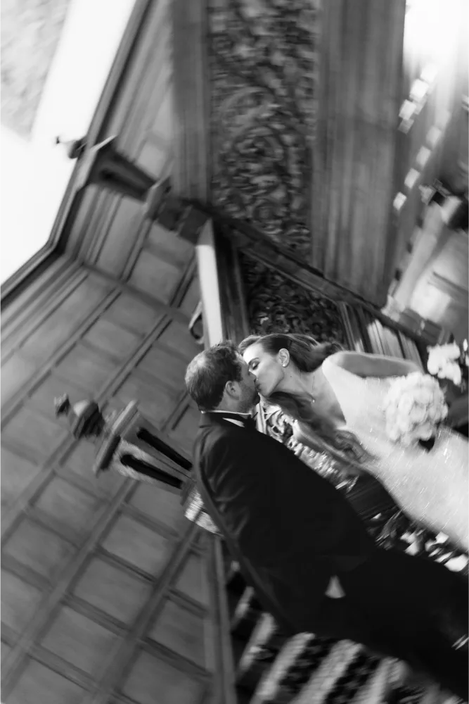 A black and white photo shows a bride and groom sharing a kiss on a wooden staircase, with the image artistically blurred and tilted, capturing a romantic and dreamy moment.