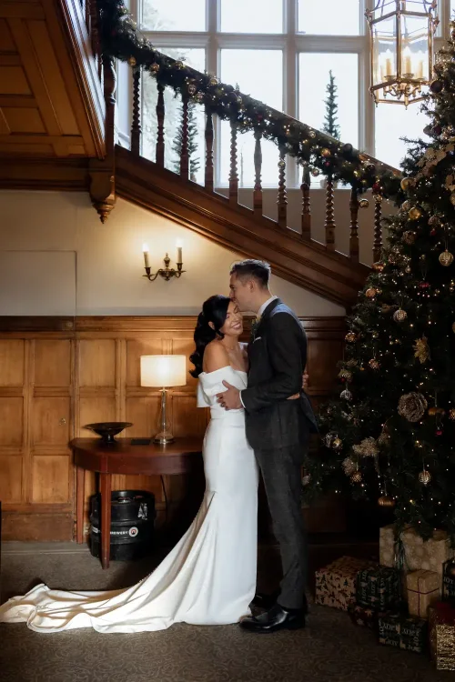 Thomas and Claire and their Elegant Winter Wedding at Hartsfield Manor, Surrey / Surrey Wedding Photographer 19 A bride and groom embrace and smile at each other by a decorated Christmas tree inside a wood-paneled room, with large windows and a staircase adorned with garland, capturing the magic of their country wedding.