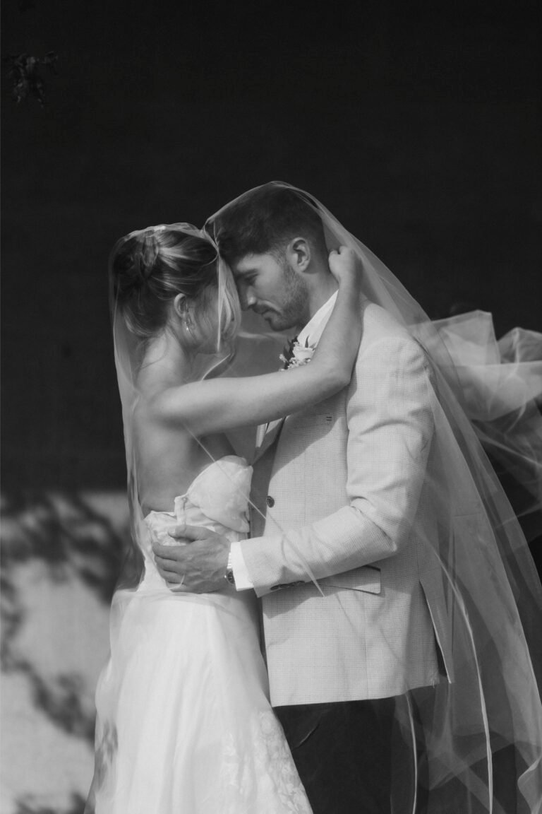 Home 4 A bride and groom embrace closely, touching foreheads as sunlight creates soft shadows around them. Captured by a London wedding photographer, their tender moment in elegant attire is framed by the bride’s flowing veil.