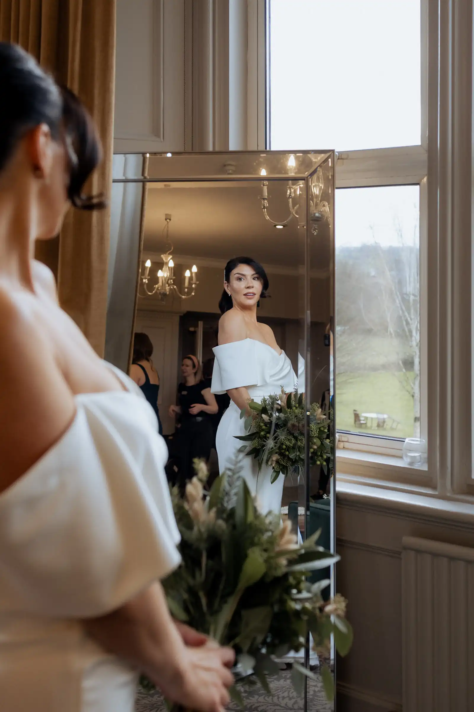 Thomas and Claire and their Elegant Winter Wedding at Hartsfield Manor, Surrey / Surrey Wedding Photographer 13 A bride in an off-shoulder white dress holds a bouquet and gazes at her reflection in a tall mirror, with large windows and a chandelier illuminating her country wedding at the farm.