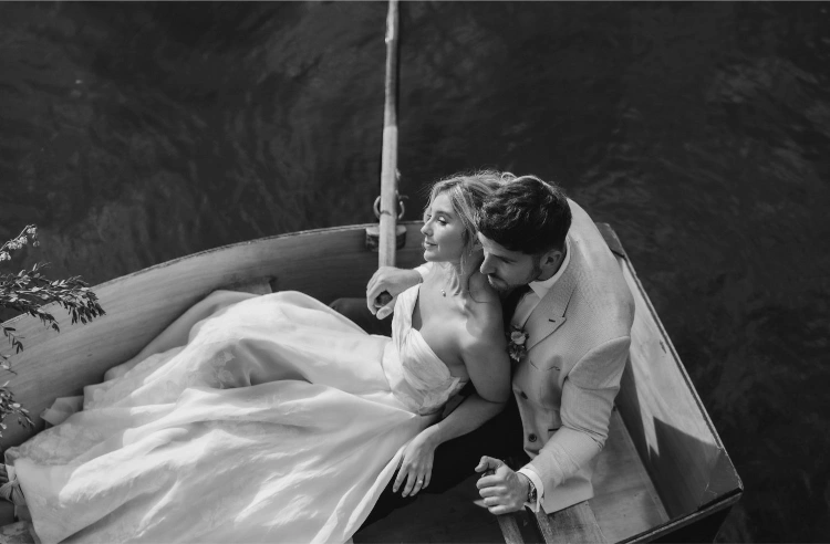 A bride and groom in wedding attire sit closely together in a small wooden rowboat on the water, embracing and looking content. The image is in black and white.