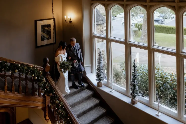 Thomas and Claire and their Elegant Winter Wedding at Hartsfield Manor, Surrey / Surrey Wedding Photographer 15 A bride in a white dress holds a bouquet while walking downstairs with an older man in formal attire during a charming country wedding. Inside the decorated venue, large windows reveal cars parked outside.