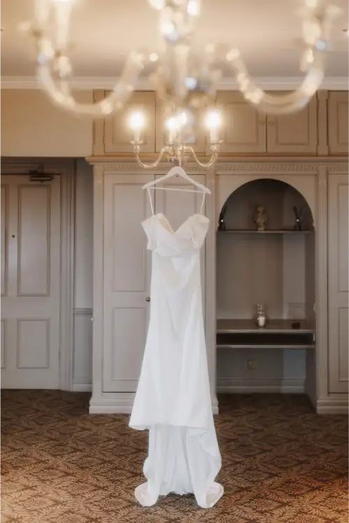 Thomas and Claire and their Elegant Winter Wedding at Hartsfield Manor, Surrey / Surrey Wedding Photographer 12 A white, elegant strapless wedding dress hangs from a chandelier in a softly lit room with beige paneled walls and patterned carpet, ready for a beautiful farm wedding in Surrey.