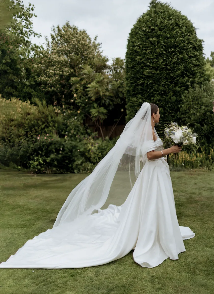 A bride in a white off-the-shoulder gown with a long train and veil stands on grass, holding a bouquet. Captured in a candid editorial style by a Surrey wedding photographer, she is surrounded by lush garden greenery.
