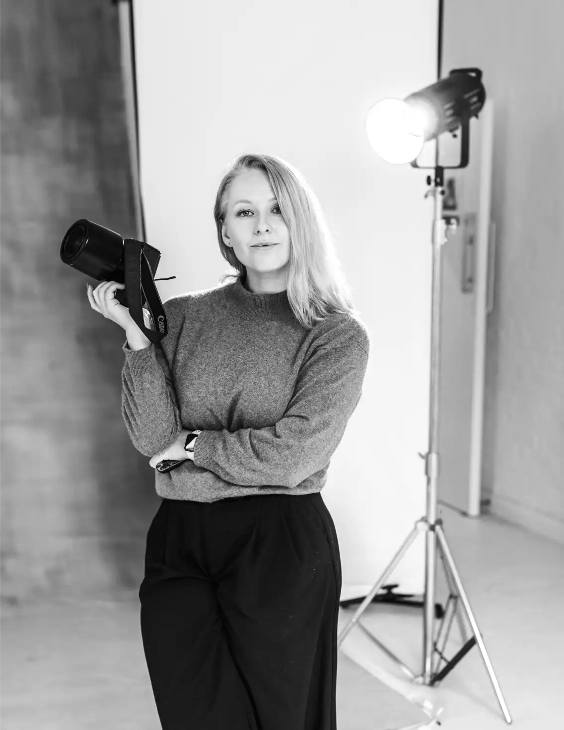 Home 3 A woman stands in a photo studio holding a camera flash, surrounded by lighting and backdrop. She wears a sweater and wide-leg pants, looking confidently at the camera—a true Surrey Wedding Photographer captured in classic black and white.