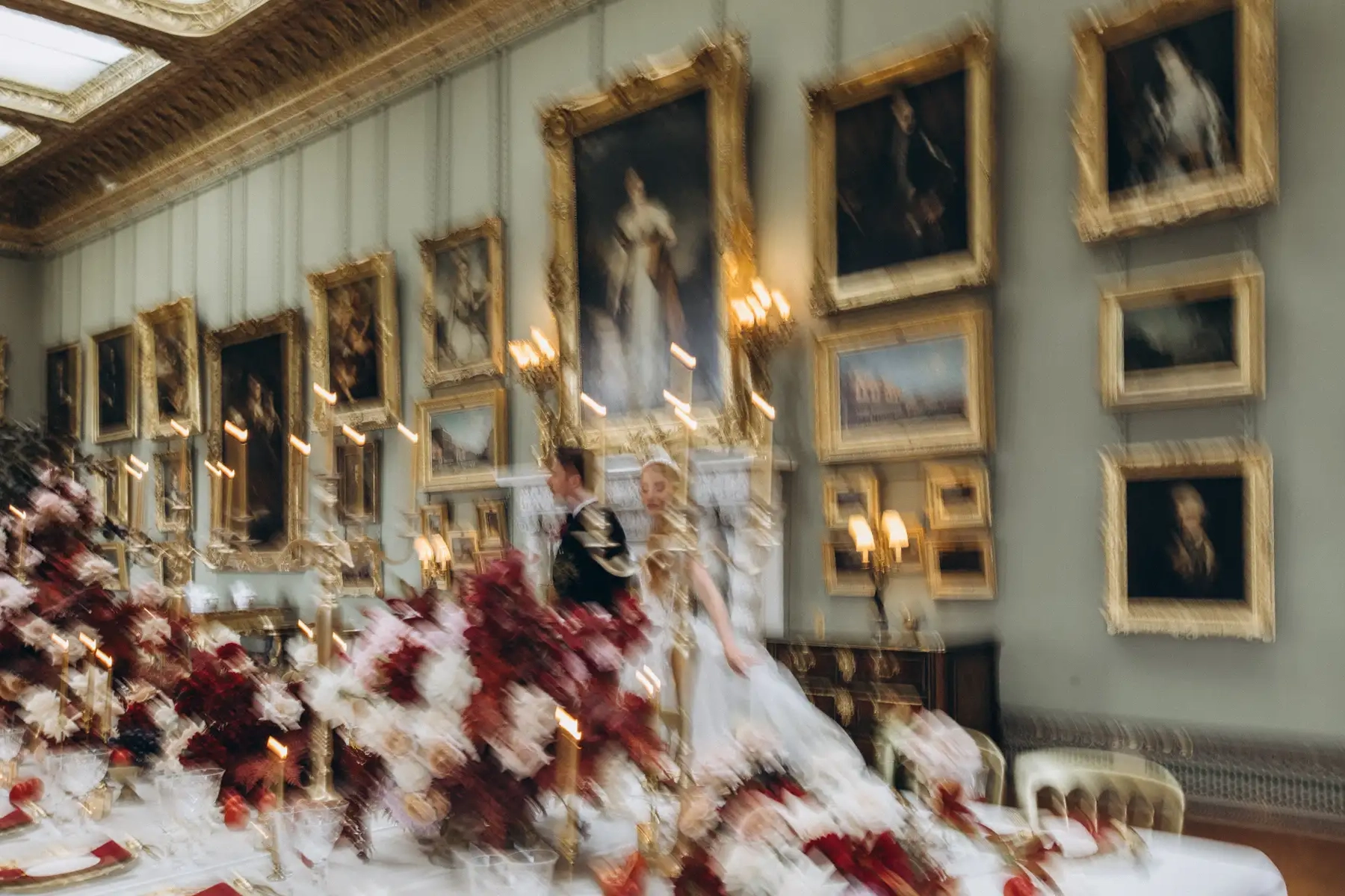 Home 2 A blurred photo of a couple standing behind an elaborate, flower-filled dining table in a grand room adorned with many framed paintings and ornate decor, captured by a Surrey Wedding Photographer known for editorial wedding photography. Photo taken by Surrey wedding photographer in Somerley House - Private Wedding Venue