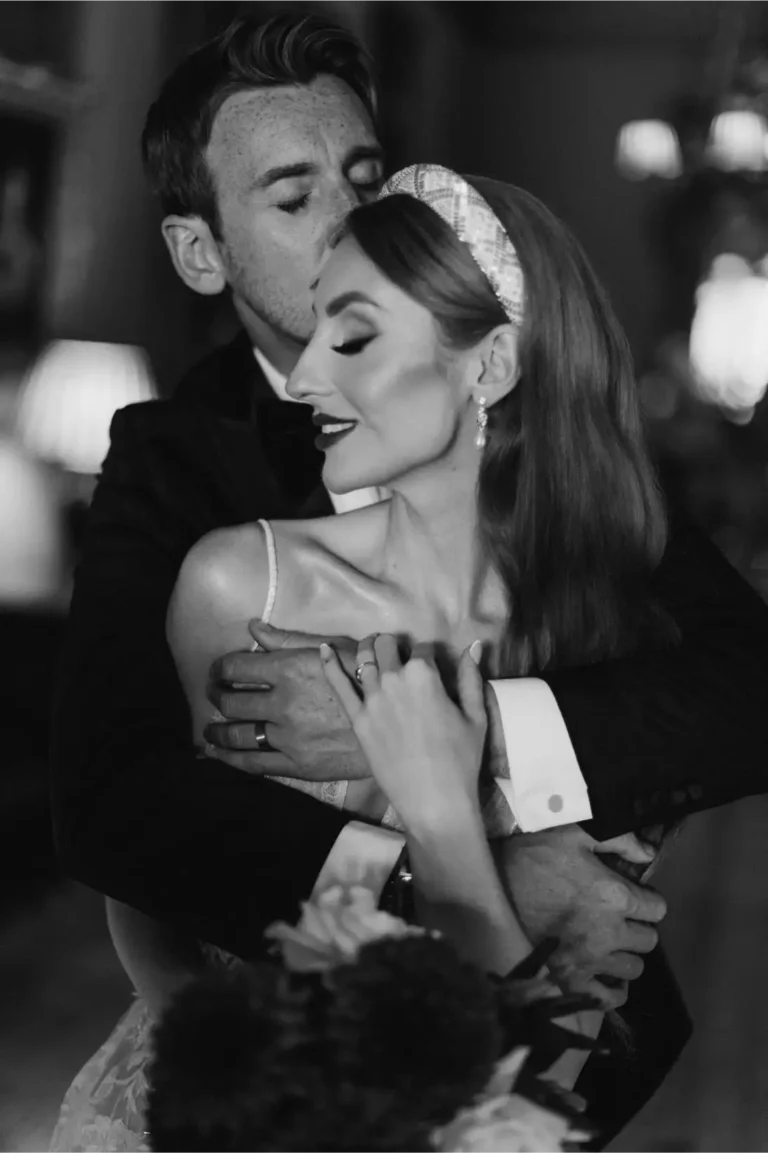 A black-and-white photo of a groom embracing his bride from behind, gently kissing her head. The bride smiles softly with eyes closed, holding his arm. They appear joyful and affectionate. Photo taken by Surrey wedding photographer in Somerley House - Private Wedding Estate