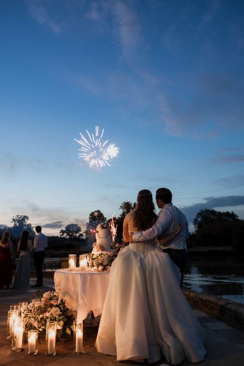 Home 2 A bride and groom stand by a candlelit, flower-adorned table watching fireworks over the lake, as guests mingle behind them—a magical moment captured by a Surrey Wedding Photographer.
