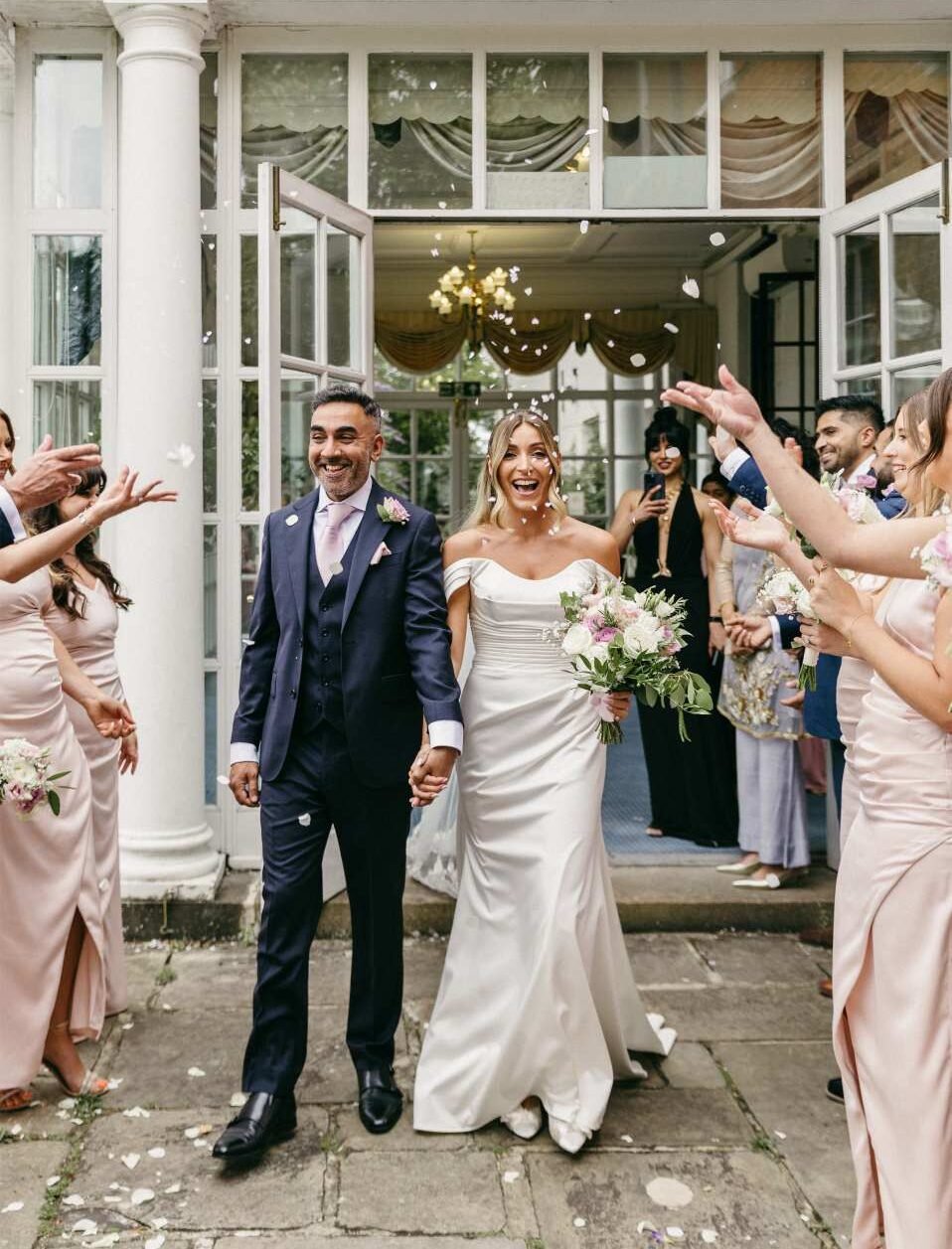 A bride and groom walk hand in hand, smiling joyfully, as wedding guests in pastel dresses throw flower petals outside a white building—captured beautifully by an editorial wedding photography London professional.