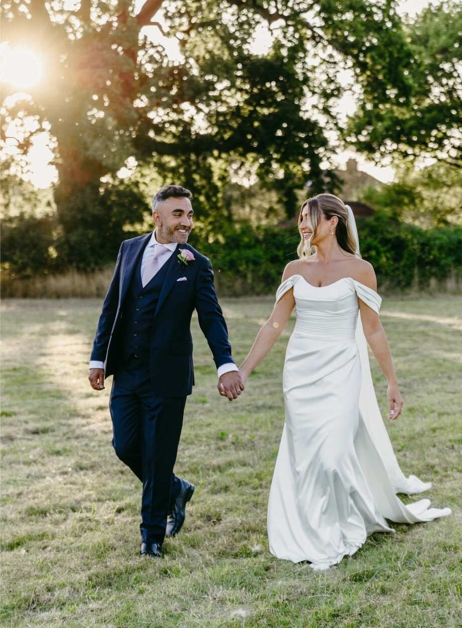 A newlywed couple holding hands and smiling at each other, walking outdoors on grass with trees and sunlight in the background—captured in elegant style by an editorial wedding photography London expert. The groom wears a suit; the bride an off-shoulder gown.