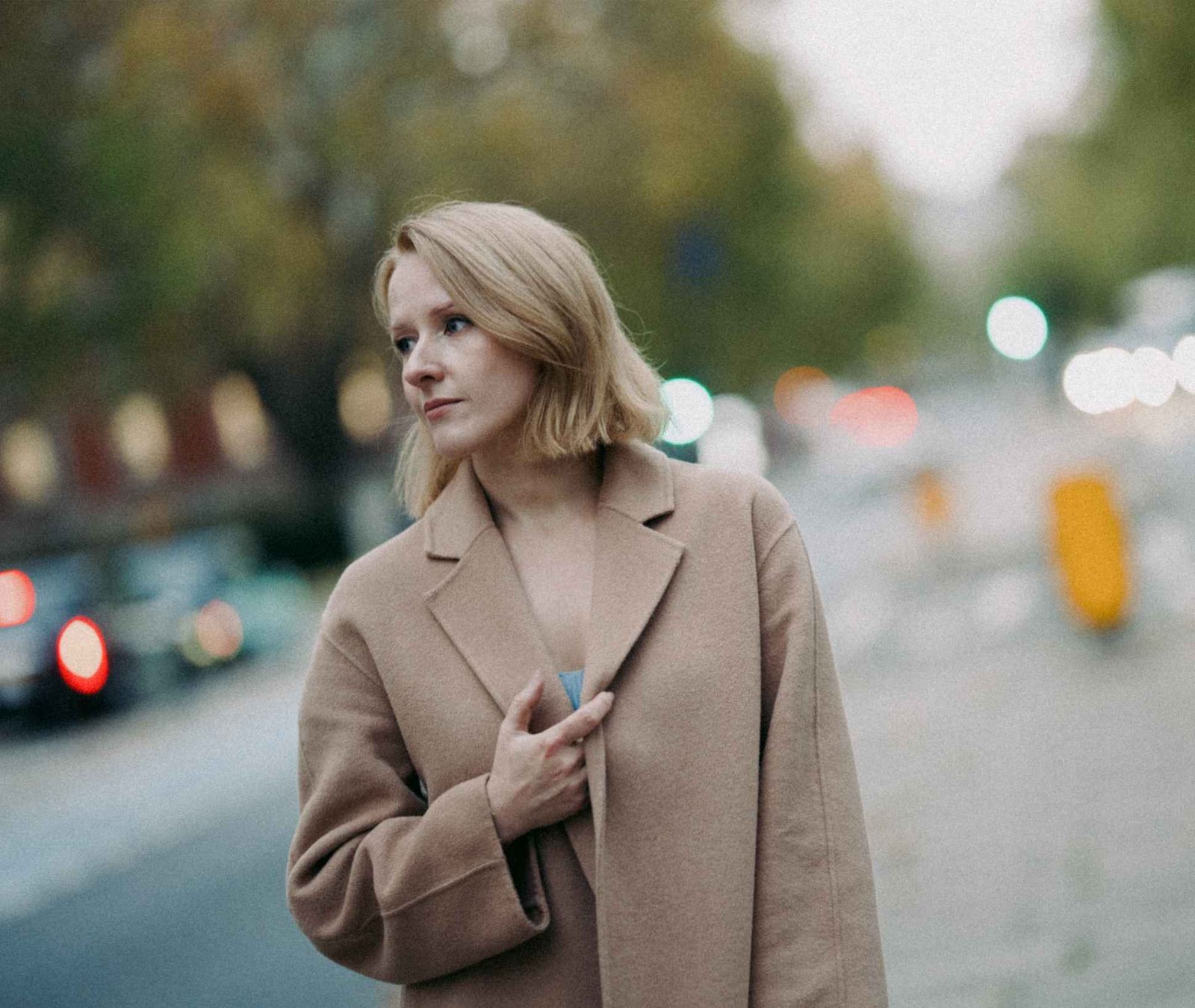 A woman with blonde hair wearing a beige coat stands outdoors near a street, looking to the side. The background is blurred with cars and trees visible, captured by a talented photographer in Surrey.