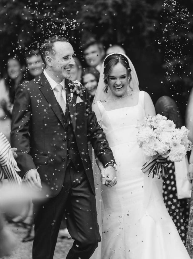 A bride and groom smile joyfully while walking hand in hand, surrounded by confetti. Captured by a Surrey Wedding Photographer, this black and white photo highlights the couple's happiness on their special day.