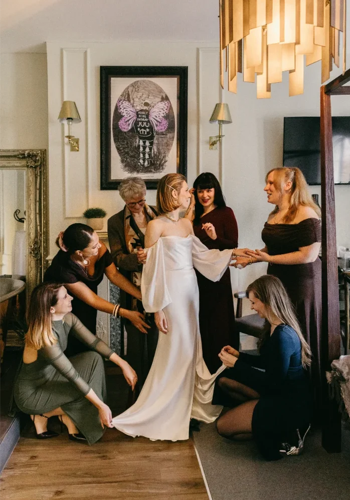 A bride in a white dress stands smiling, surrounded by women helping with her gown and accessories in a warmly lit room—a joyful moment beautifully captured by a Surrey Wedding Photographer before the ceremony. London wedding photographed by Dina Ward Photography