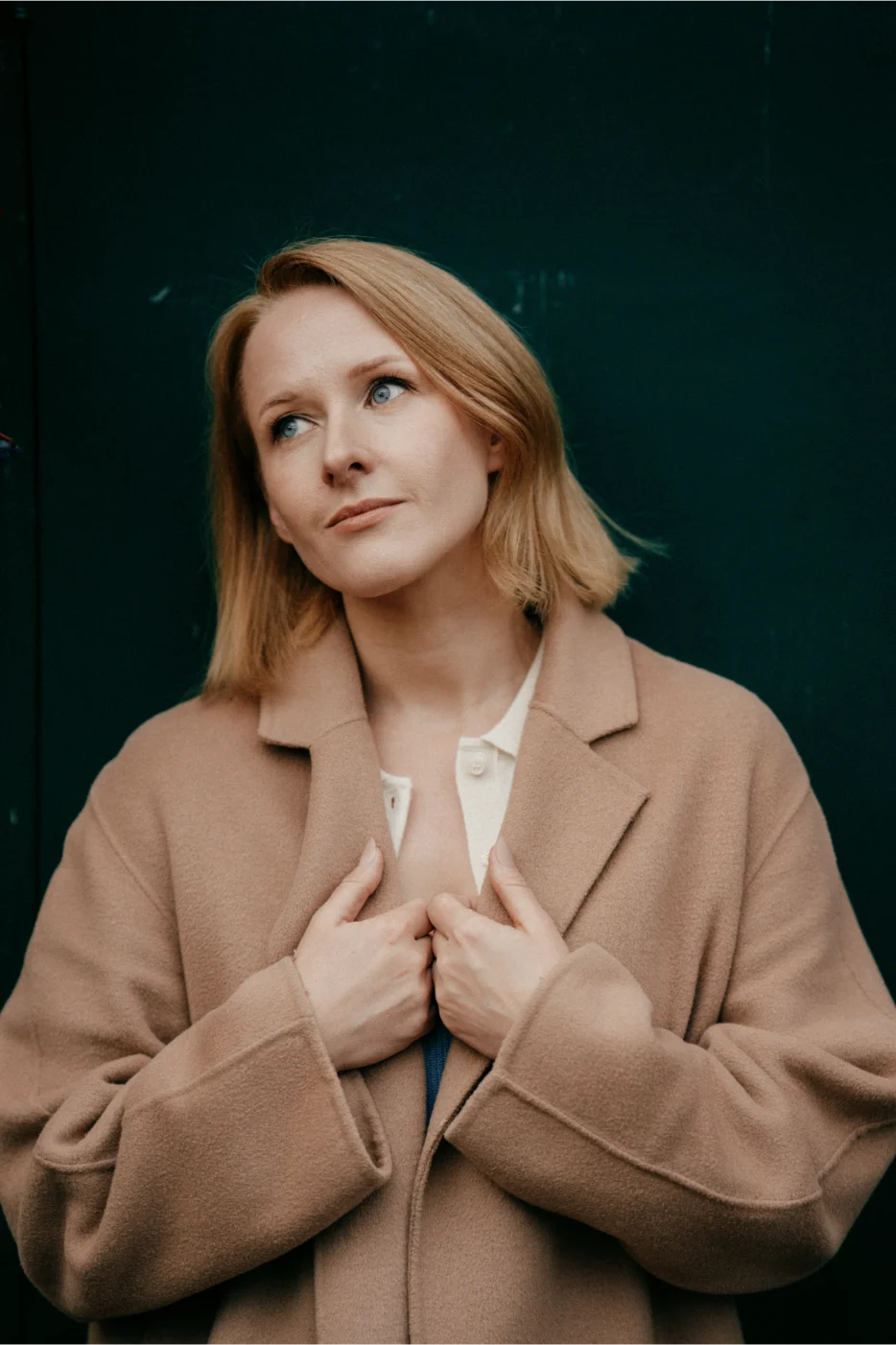 A woman with blonde hair wearing a tan coat stands against a dark green background, looking slightly upward with a thoughtful expression—her pose reminiscent of an editorial by a London wedding photographer.