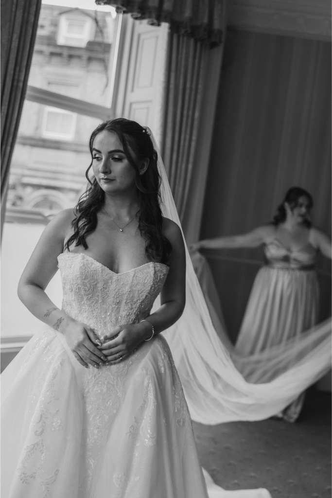 A bride in a strapless wedding dress stands near a window, looking serene. Behind her, a bridesmaid holds the bride’s long veil. Captured in black and white by a top newborn photographer known for timeless moments.