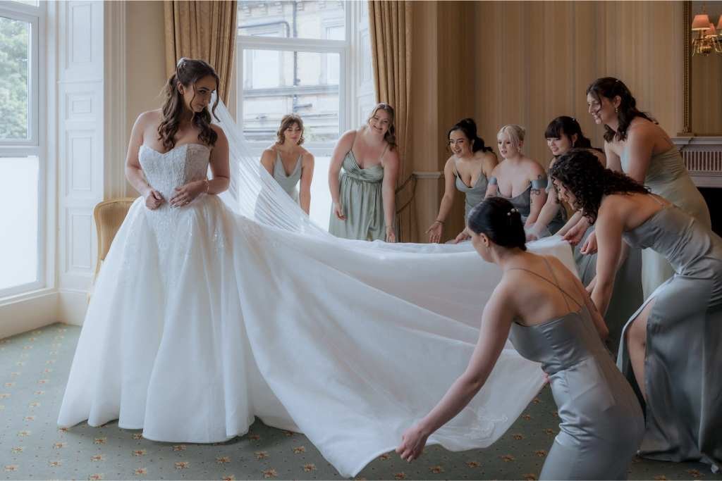 A bride in a white wedding dress stands by a window as seven bridesmaids in light gray dresses adjust her long train in an elegant, softly lit room—captured with the artistry of a top newborn photographer.
