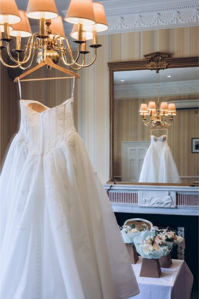 A white wedding dress hangs from a chandelier in a classic room with striped wallpaper, a large mirror, and a mantel decorated with pastel floral bouquets—captured beautifully by a top newborn photographer. The dress and chandelier are reflected in the mirror.